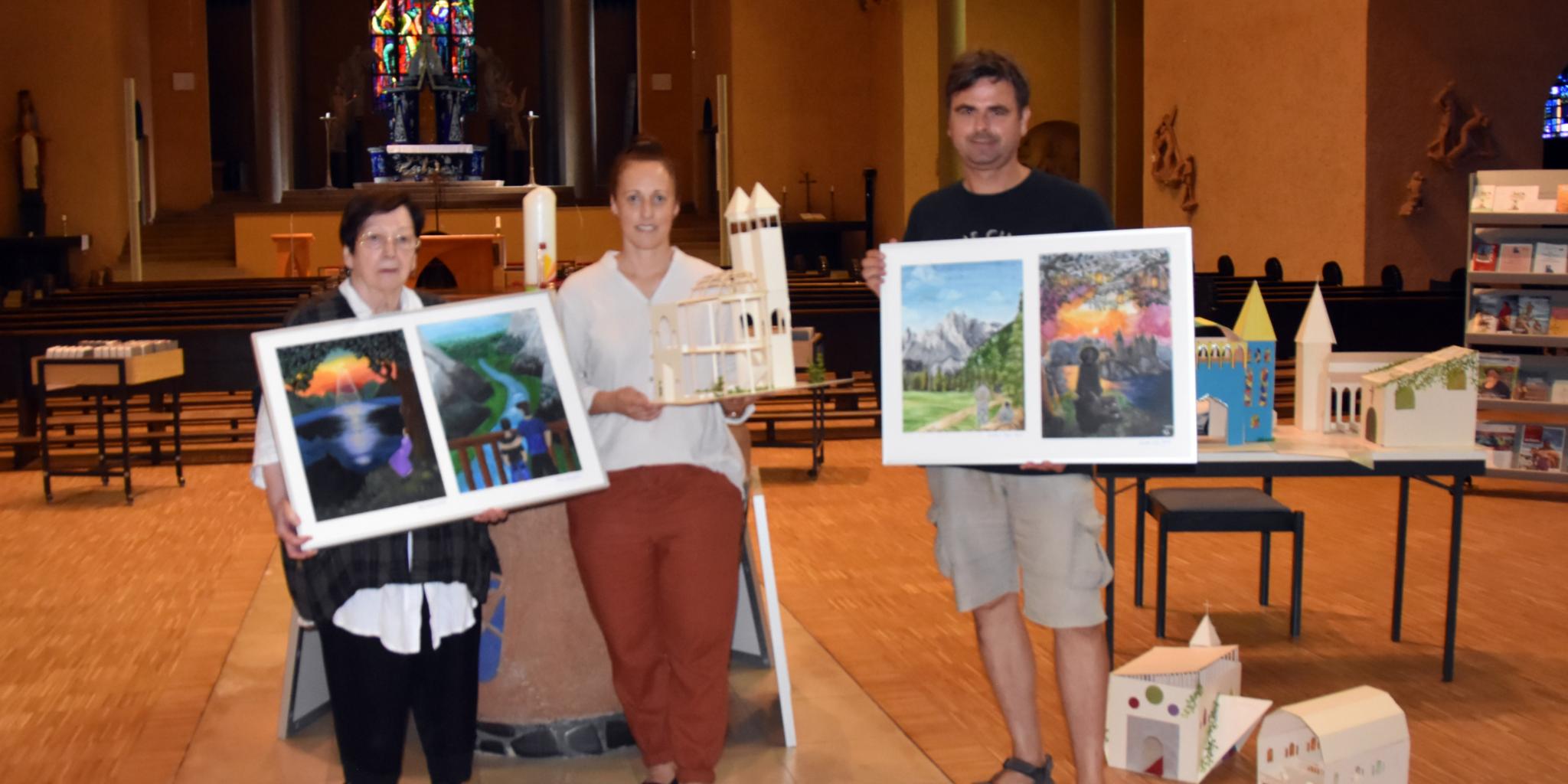 Man sieht drei Personen in einer Kirche stehen. Alle halten ein Kunstwerk der Schülerinnen und Schüler in der Hand.