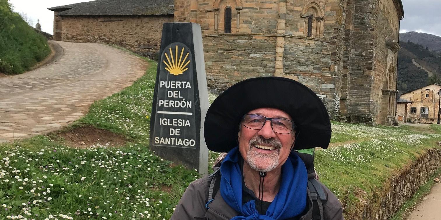 Ein älterer Mann mit Brille und großem Sonnenhut steht lächelnd vor einer alten romanischen Kirche aus Stein mit halbrunder Apsis. Neben ihm steht ein Wegweiser mit der Aufschrift „Puerta del Perdón – Iglesia de Santiago“, was auf den Jakobsweg hinweist. Im Hintergrund führen ein gepflasterter Weg und eine Wiese mit Gänseblümchen zur Kirche. (Alt-Text erstellt mit Unterstützung von ChatGPT)