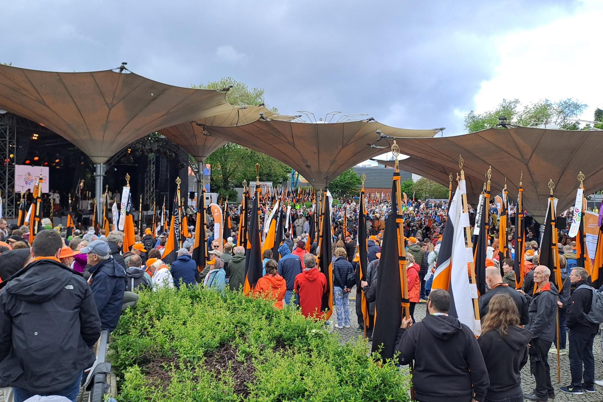 Auf dem Bild ist eine große Menschenmenge bei einer Veranstaltung im Freien zu sehen, viele von ihnen tragen orangefarbene Kleidung oder Accessoires. Zahlreiche orange-schwarze Fahnen mit goldenen Spitzen werden aufrecht gehalten. Im Hintergrund befindet sich eine überdachte Bühne mit Lautsprechern und Bildschirmen unter großen, zeltartigen Dächern. (Alt-Text erstellt mit Unterstützung von ChatGPT)