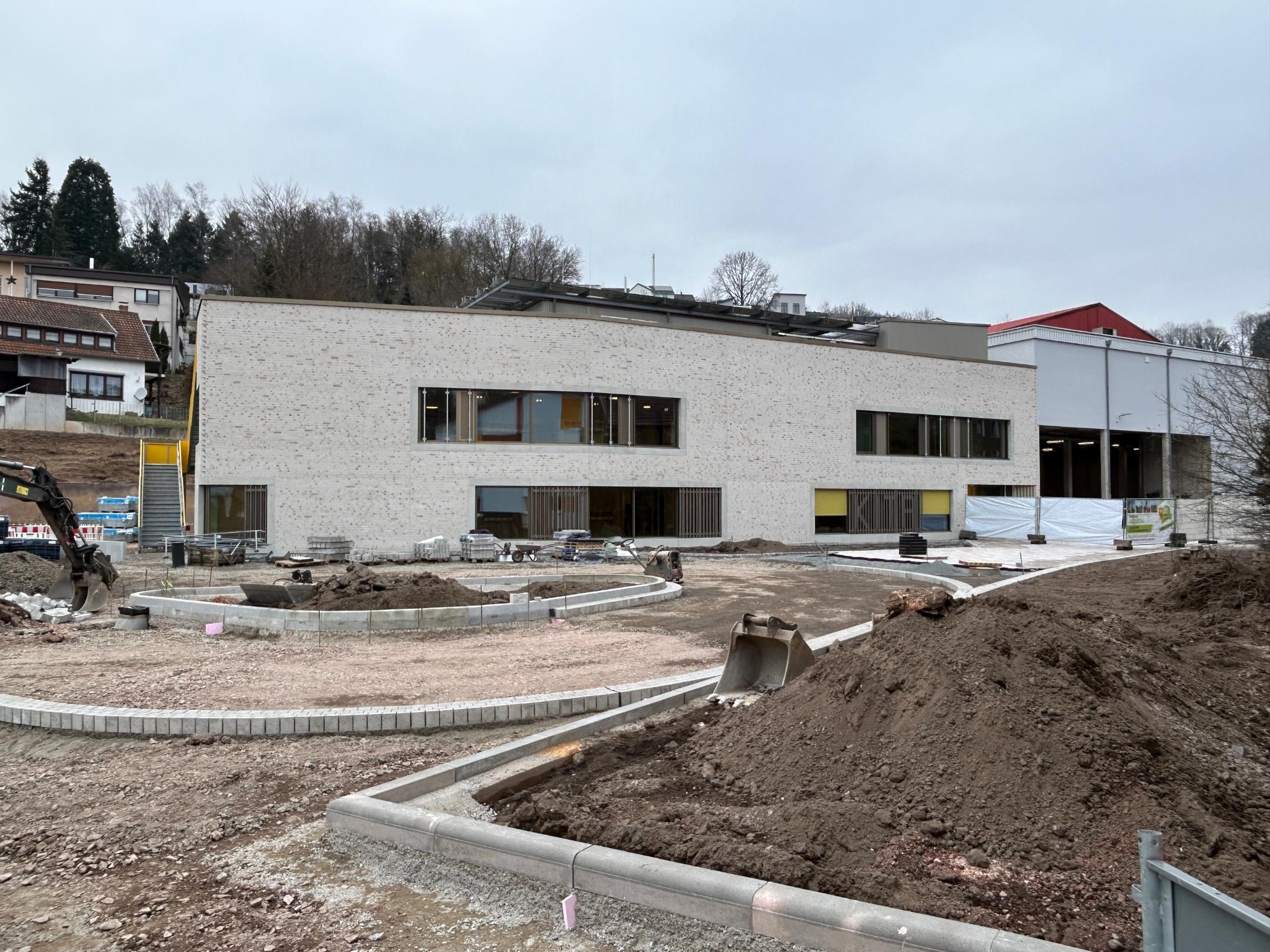 Auf einer Baufläche steht ein hellgraues Gebäude, im Außenbereich wird noch gearbeitet