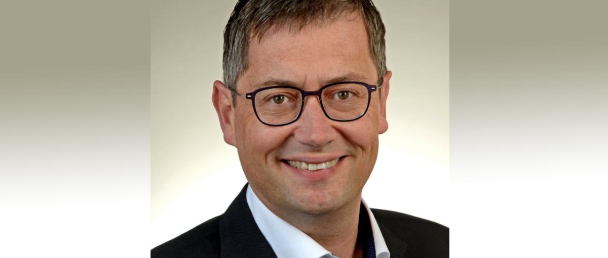 Ein Mann, mittleren Alters, mit Brille und kurzen Haaren, lächelt in die Kamera. Er trägt einen schwarzen Anzug ohne Krawatte