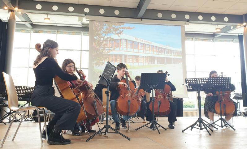 Cellisten und Cellistinnen mit ihren Instrumenten vor einer Leinwand mit der Aufschrift 'Willi-Graf-Gymnasium'