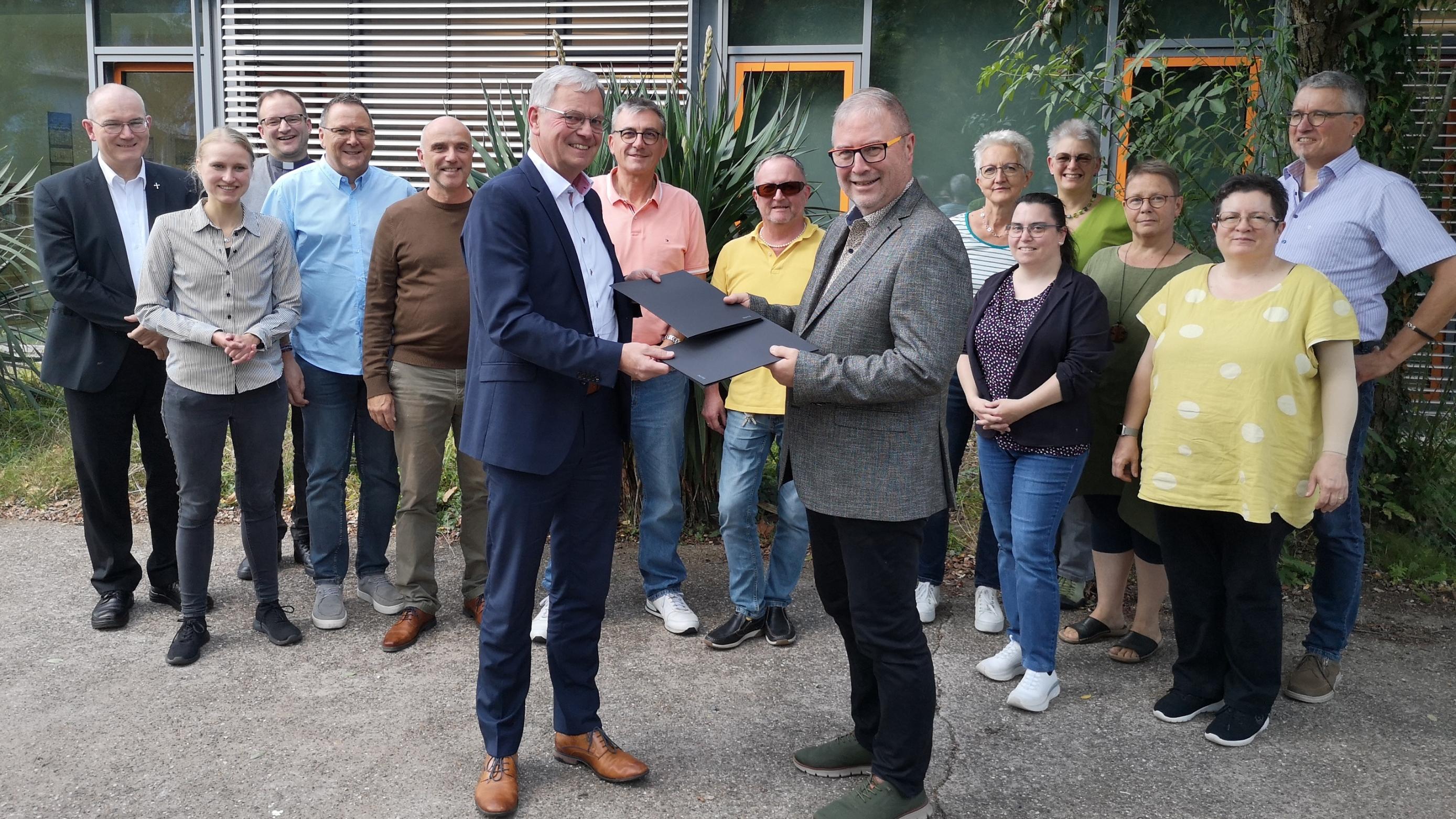 Generalvikar Dr. Ulrich Graf von Plettenberg (v.l.) und Superintendent Christian Weyer (v.r.) nehmen wechselseitig die unterzeichnete Kooperationsvereinbarung zur Krankenhausseelsorge in Empfang. Dahinter Vertretende der Klinikseelsorgenden im Saarland