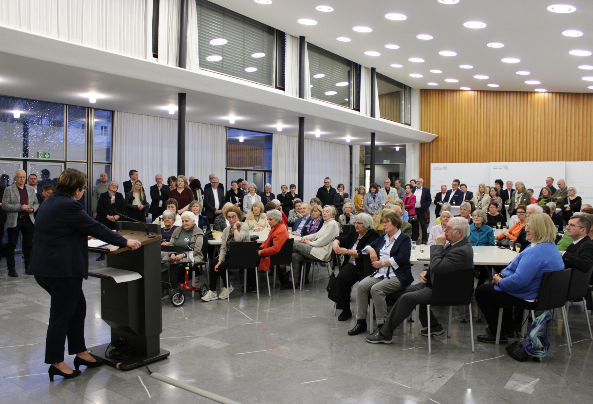 Eine Frau in dunklem Anzug steht in einem Saal am Rednerpult und spricht zu den Gästen