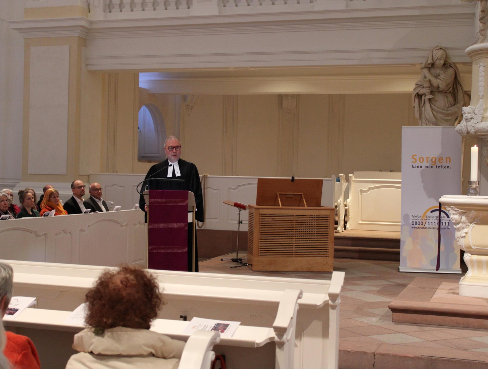 Ein Mann in dunklem Gewand steht an einem Pult neben dem Altar und spricht zu den Gästen in der Kirche