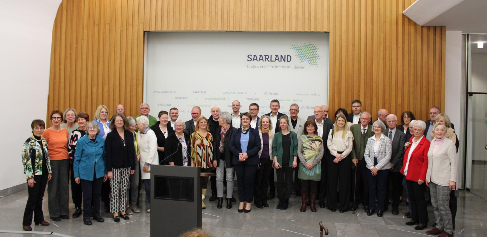 Eine Gruppe von mehr als zwanzig Menschen steht vor einer Wand aus Holz, im Hintergrund ist auf einem Plakat Saarland zu lesen