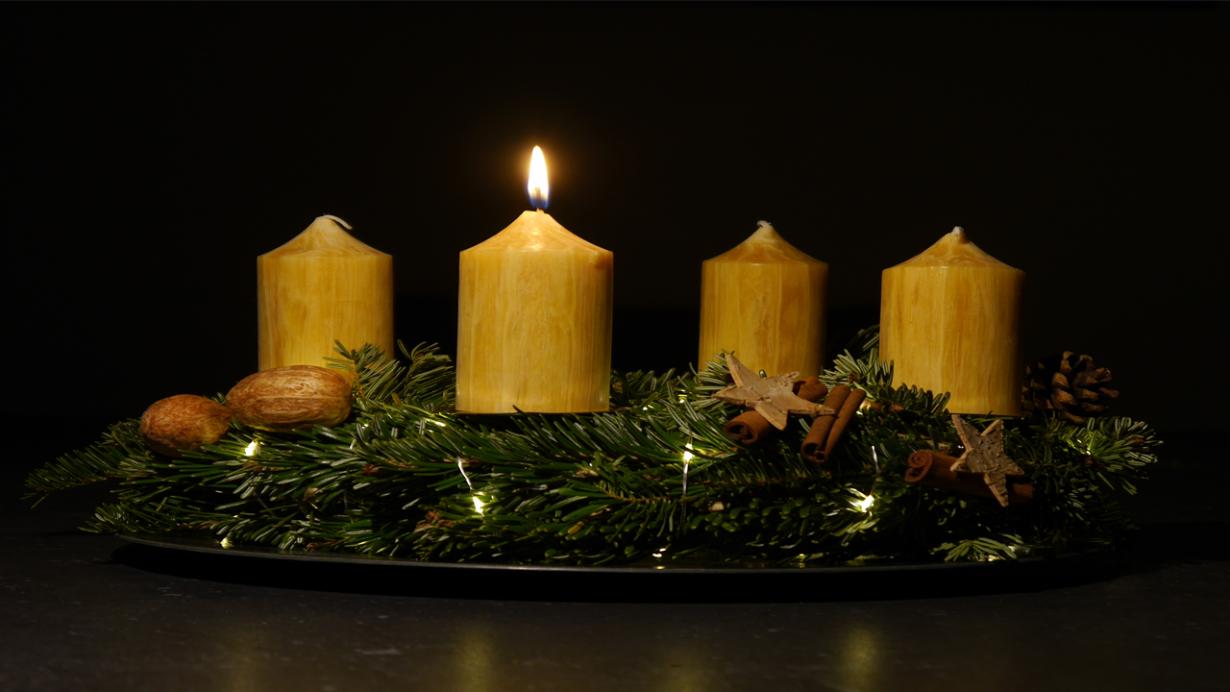 Das Foto zeigt in Großaufnahme einen Adventskranz vor dunklem Hintergrund. Der Adventskranz hat honigfarbene Kerzen, von denen eine brennt.