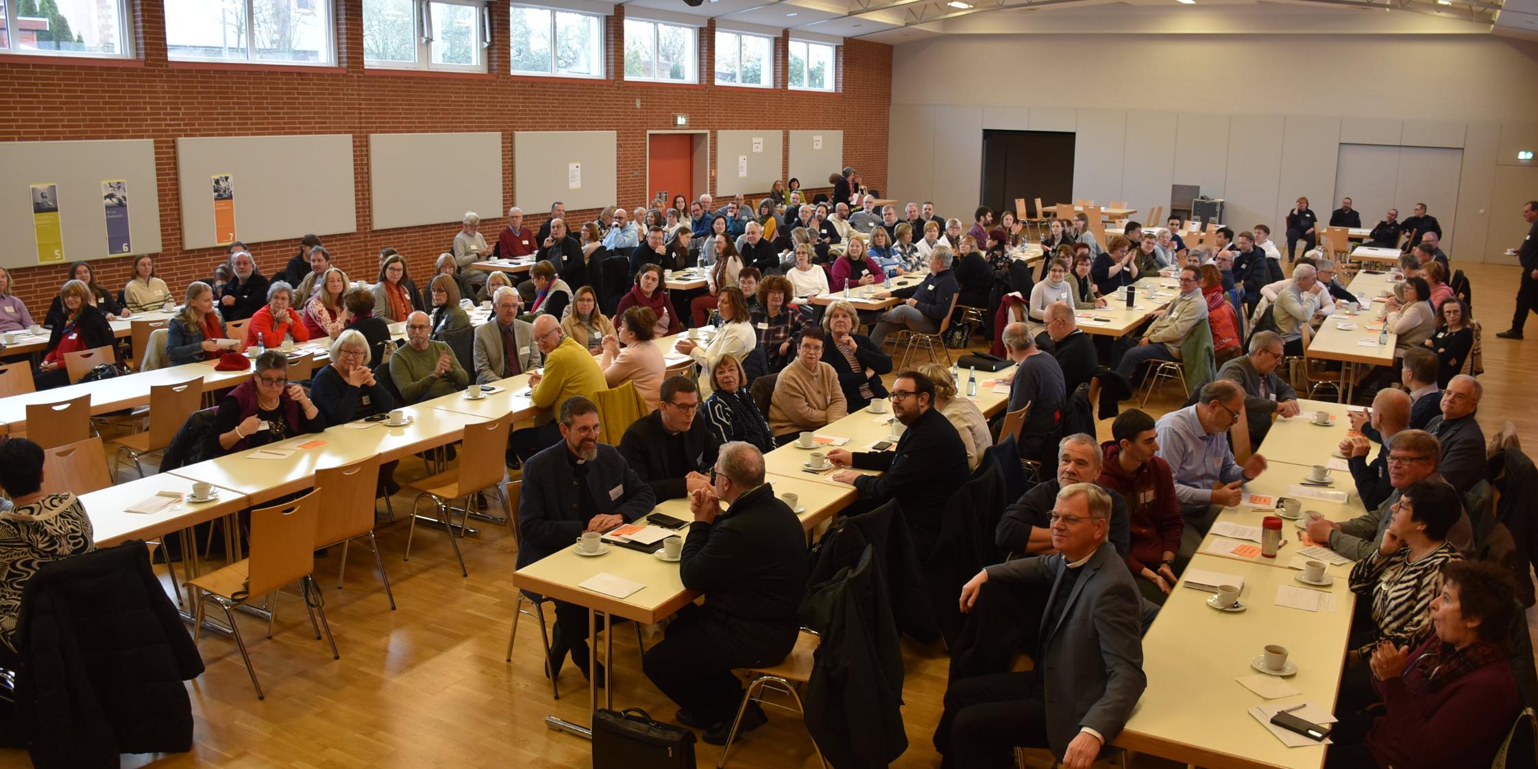 In einem großen Raum sitzen viele Menschen an langen Tischen