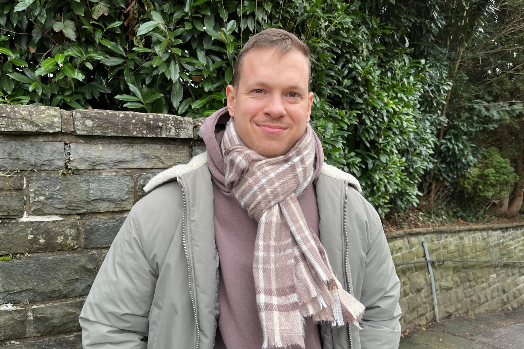 Ein junger Mann mit kurzem, hellbraunem Haar und freundlichem Lächeln steht vor einer Natursteinmauer mit dichten grünen Büschen im Hintergrund. Er trägt eine olivgrüne Winterjacke mit heller Kapuze, einen braunen Kapuzenpullover und einen beigefarbenen karierten Schal, der um seinen Hals gewickelt ist. Seine Hände sind in den Jackentaschen, und er blickt direkt in die Kamera. (Alt-Text erstellt mit Unterstützung von ChatGPT)