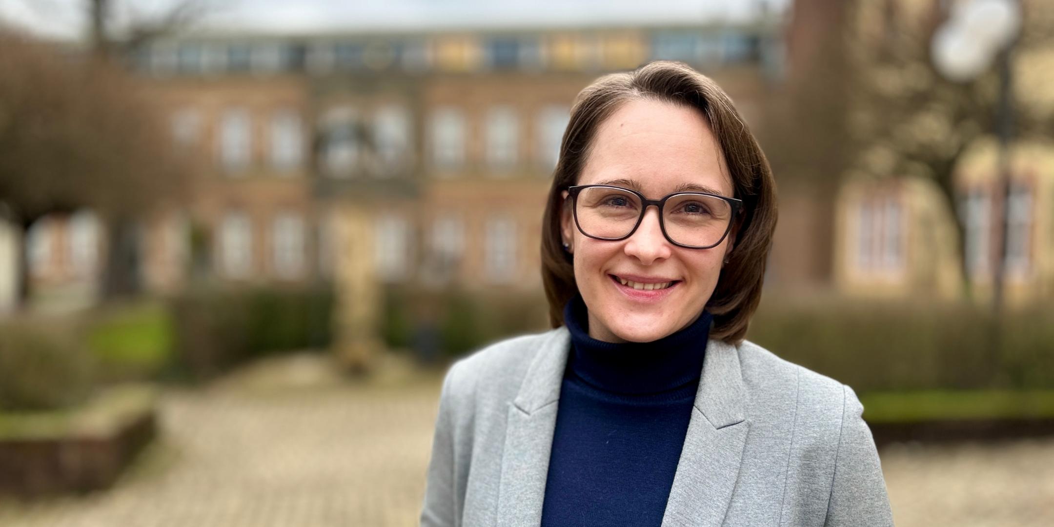 Eine Frau mit schulterlangem braunen Haar und Brille steht lächelnd im Freien vor einem historischen Backsteingebäude. Sie trägt einen grauen Blazer über einem dunkelblauen Rollkragenpullover. Der Hintergrund ist leicht unscharf, mit einem gepflasterten Weg, Hecken und Bäumen.