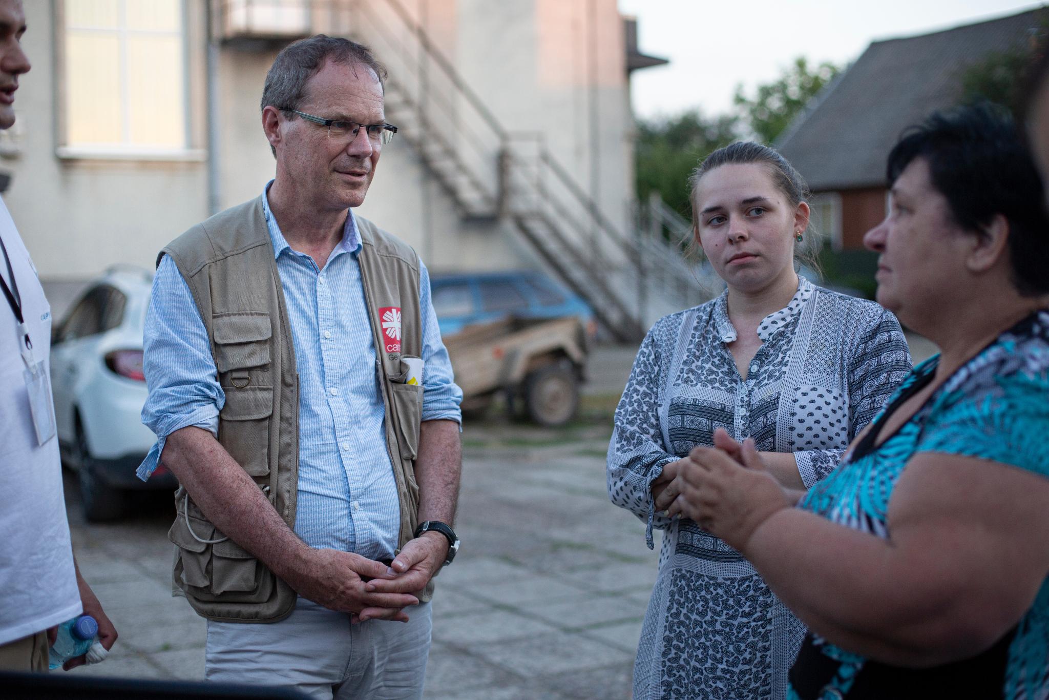 Ein Mann mit Brille und Weste mit dem Logo von Caritas international (Dr. Oliver Müller) spricht mit zwei Frauen im Freien. Eine junge Frau mit gemustertem Kleid hört aufmerksam zu, während die andere Frau in Blau spricht. Im Hintergrund sind ein Auto, ein Anhänger und ein Gebäude mit einer Außentreppe zu sehen. (Alt-Text erstellt mit ChatGPT)