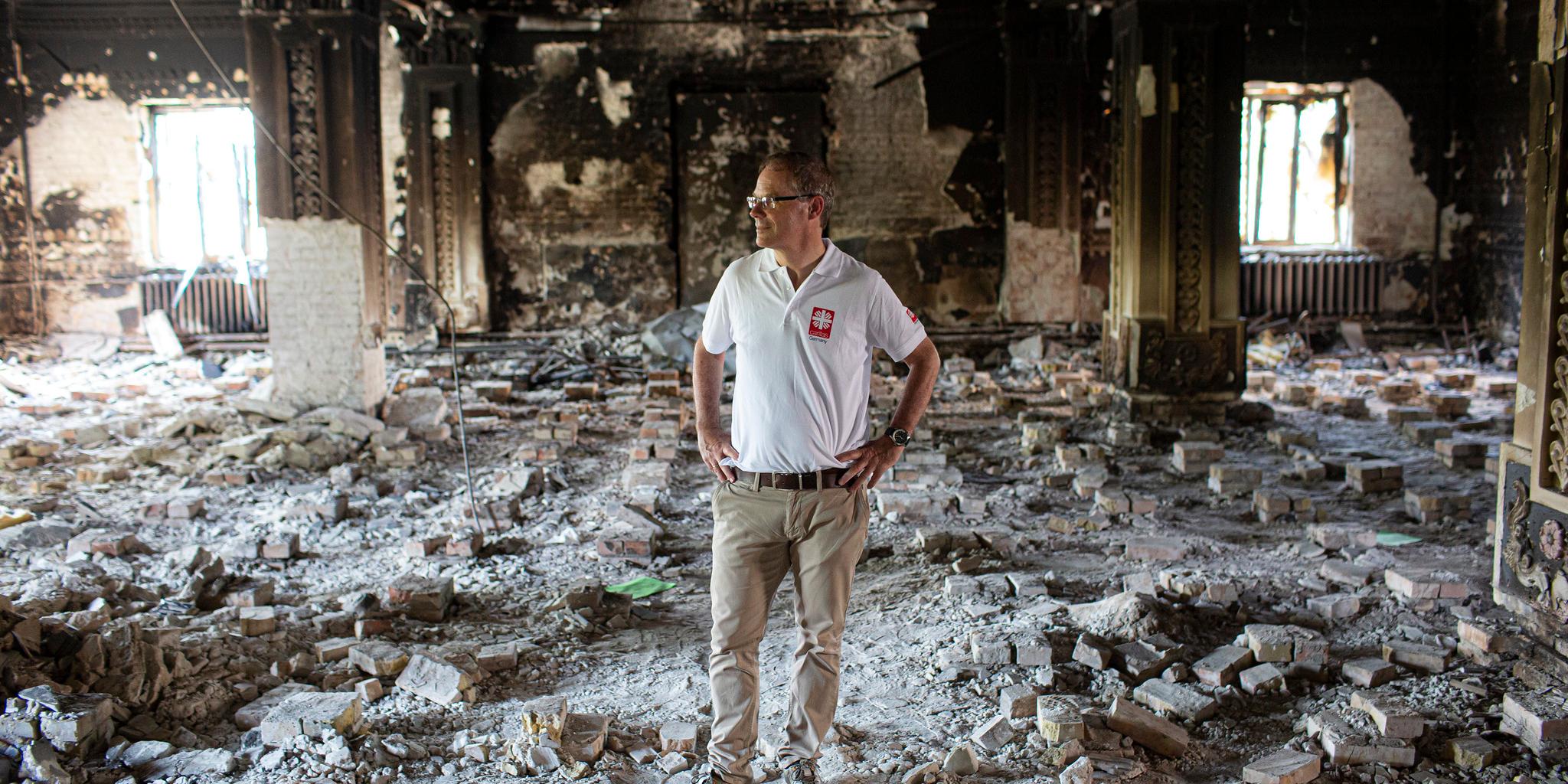 Ein Mann mit Brille und einem weißen Polohemd mit dem Logo von Caritas international (Dr. Oliver Müller) steht mit in die Hüften gestützten Händen in den Trümmern eines offenbar ausgebrannten Gebäudes. Der Boden ist mit Schutt und Ziegelsteinen bedeckt, die Wände und Säulen sind stark beschädigt. Im Hintergrund sind verkohlte Wände, zwei Fenster und ein Heizkörper zu sehen. (Alt-Text erstellt mit ChatGPT)