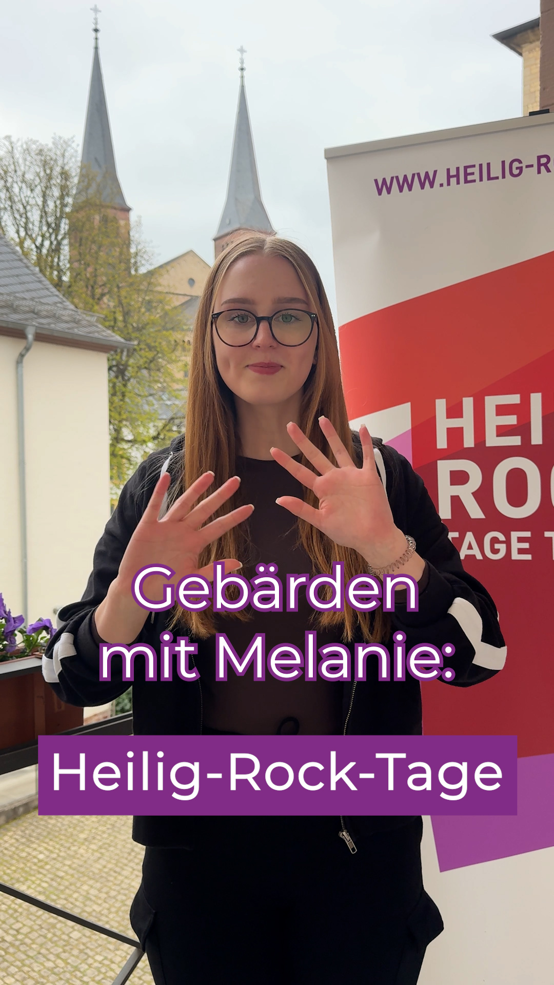 Auszubildende macht Gebärden zum Thema Heilig-Rock-Tage und steht vor einem Plakat der Heilig-Rock-Tage.
