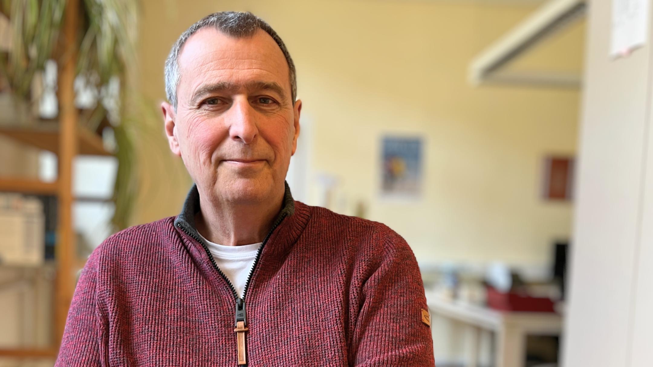 Ein älterer Mann (es handelt sich um Hubertus Kesselheim) mit kurzen grauen Haaren und freundlichem Gesichtsausdruck sitzt in einem Büro und schaut direkt in die Kamera. Er trägt einen rotbraunen Strickpullover mit Reißverschluss über einem weißen T-Shirt. Im Hintergrund sind unscharf ein Schreibtisch, Pflanzen und Büromaterialien zu sehen. (Alt-Text erstellt mit Unterstützung von KI)