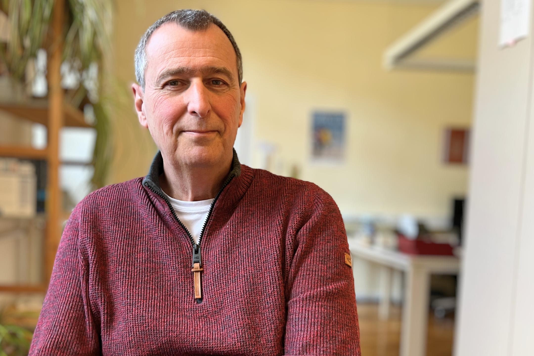 Ein älterer Mann (es handelt sich um Hubertus Kesselheim) mit kurzen grauen Haaren und freundlichem Gesichtsausdruck sitzt in einem Büro und schaut direkt in die Kamera. Er trägt einen rotbraunen Strickpullover mit Reißverschluss über einem weißen T-Shirt. Im Hintergrund sind unscharf ein Schreibtisch, Pflanzen und Büromaterialien zu sehen. (Alt-Text erstellt mit Unterstützung von KI)