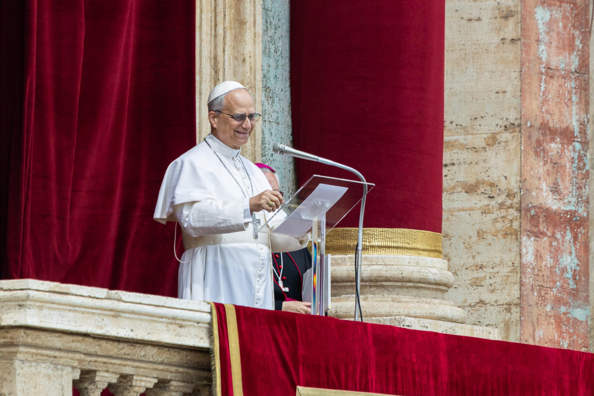 Papst Leo XIV. steht in weißer päpstlicher Kleidung auf der Loggia des Petersdoms vor einem roten Samtvorhang und spricht an einem Mikrofon. Er trägt ein Brustkreuz und eine weiße Kappe (Zucchetto) und lächelt. Eine weitere geistliche Person in dunkler Kleidung ist teilweise hinter ihm zu sehen.