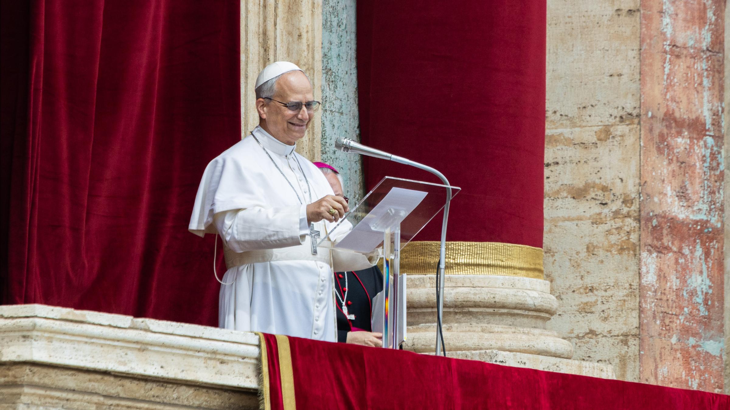 Papst Leo XIV. steht in weißer päpstlicher Kleidung auf der Loggia des Petersdoms vor einem roten Samtvorhang und spricht an einem Mikrofon. Er trägt ein Brustkreuz und eine weiße Kappe (Zucchetto) und lächelt. Eine weitere geistliche Person in dunkler Kleidung ist teilweise hinter ihm zu sehen.