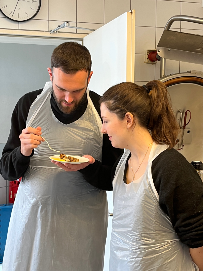 Ein junger Mann und eine junge Frau in Schürze schauen auf einen kleinen Teller mit Essen.