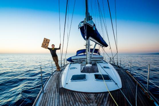 Joshi Nichell steht auf einem weißen Segelboot, das sich auf offener See befindet. Mit einer Hand hält er sich am Segel fest und lehnt sich über das Boot hinaus; mit der anderen Hand hält er ein Schild fest.