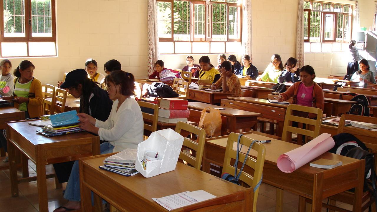 Das Foto zeigt einen Klassenraum in Bolivien. Holztische mit jeweils zwei Stühlen stehen in drei Reihen nebeneinander. Auf den Tischen in der vorderen Reihen liegen Bücher und Papier, hier sitzen keine Schülerinnen und Schüler. Die Tische in der mittleren und hinteren Reihe sind mit jeweils ein oder zwei Schülerinnen besetzt, die schreiben, lesen und sich unterhalten. Die hintere Reihe steht vor einer Wand mit Fenstern, draußen scheint die Sonne, vor den Fenstern stehen Büsche oder eine Hecke.