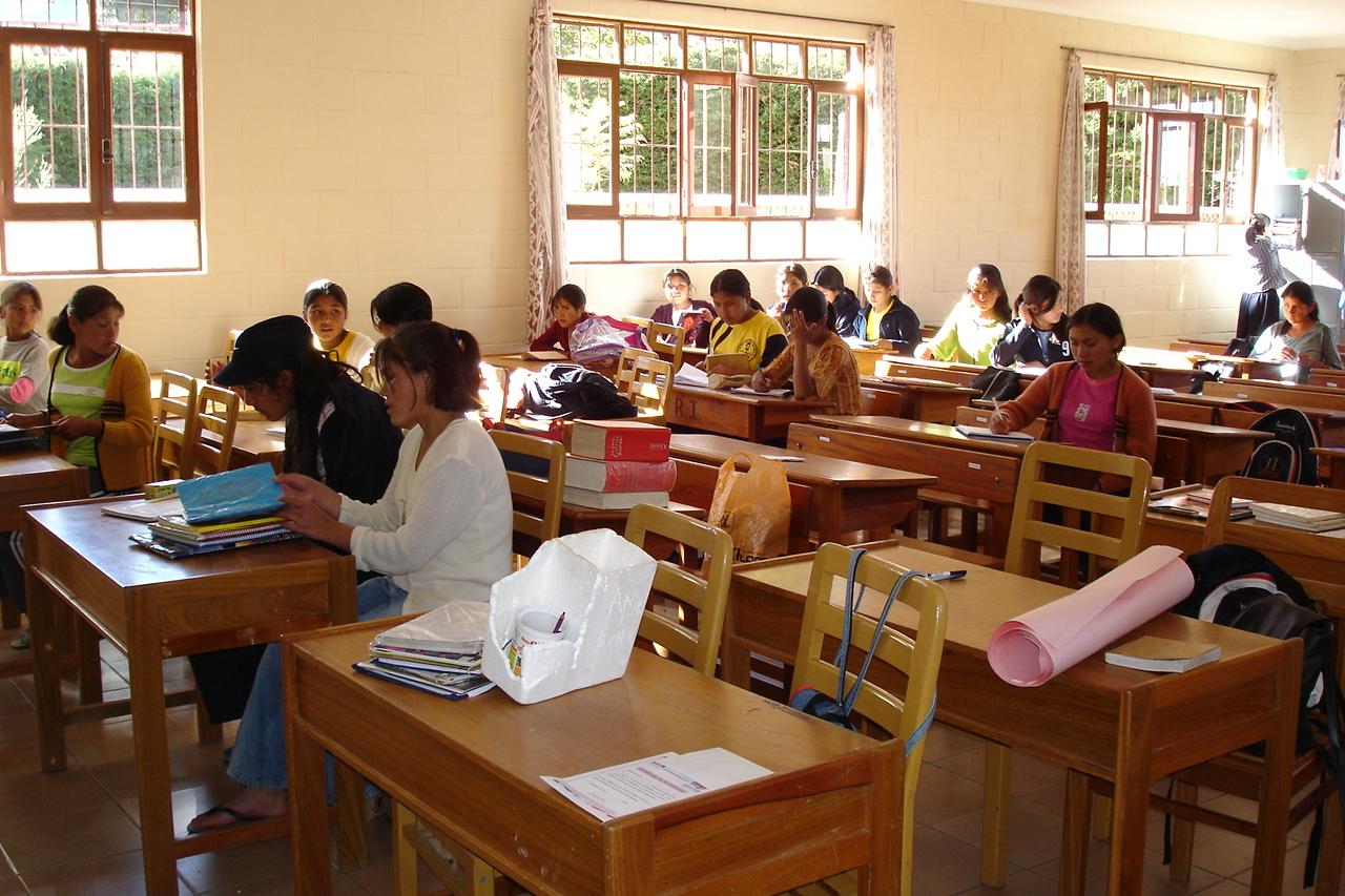 Das Foto zeigt einen Klassenraum in Bolivien. Holztische mit jeweils zwei Stühlen stehen in drei Reihen nebeneinander. Auf den Tischen in der vorderen Reihen liegen Bücher und Papier, hier sitzen keine Schülerinnen und Schüler. Die Tische in der mittleren und hinteren Reihe sind mit jeweils ein oder zwei Schülerinnen besetzt, die schreiben, lesen und sich unterhalten. Die hintere Reihe steht vor einer Wand mit Fenstern, draußen scheint die Sonne, vor den Fenstern stehen Büsche oder eine Hecke.