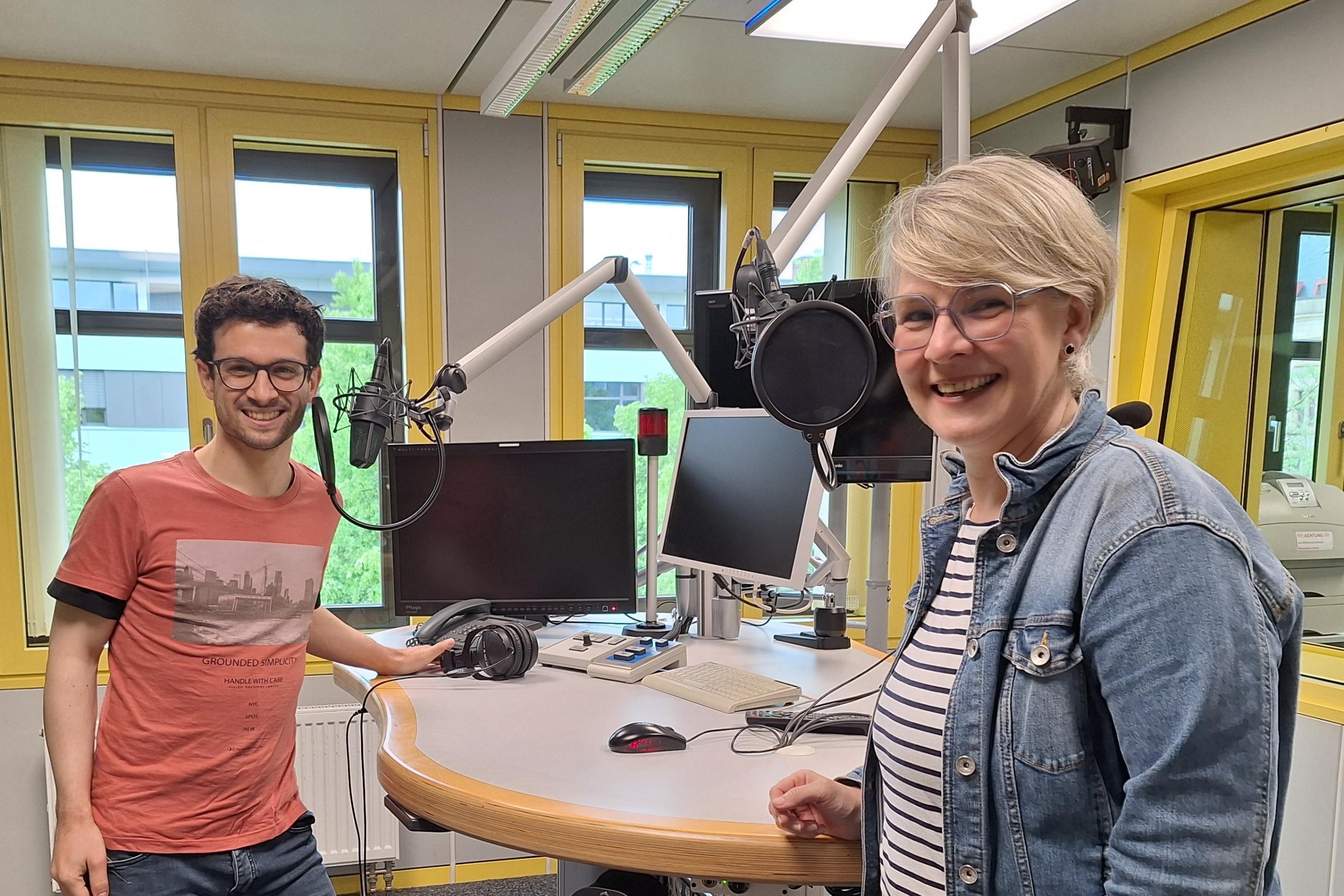 Das Foto zeigt Christopher Hoffmann (links) und Nicole Stockschlaeder (rechts). Sie stehen in einem Hörfunkstudio an einem Tisch mit Mikrofonen und lächeln freundlich in die Kamera.