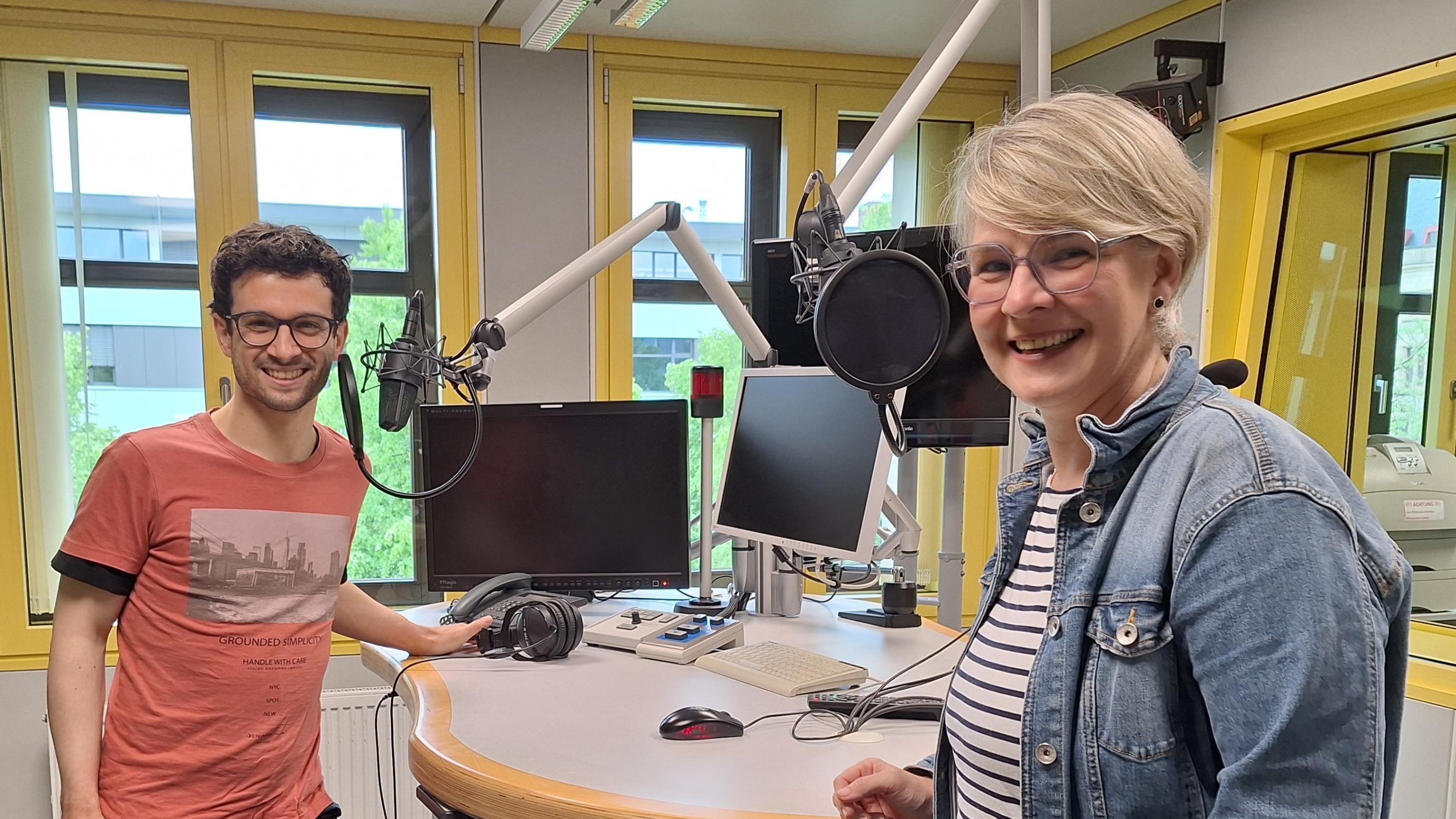 Das Foto zeigt Christopher Hoffmann (links) und Nicole Stockschlaeder (rechts). Sie stehen in einem Hörfunkstudio an einem Tisch mit Mikrofonen und lächeln freundlich in die Kamera.