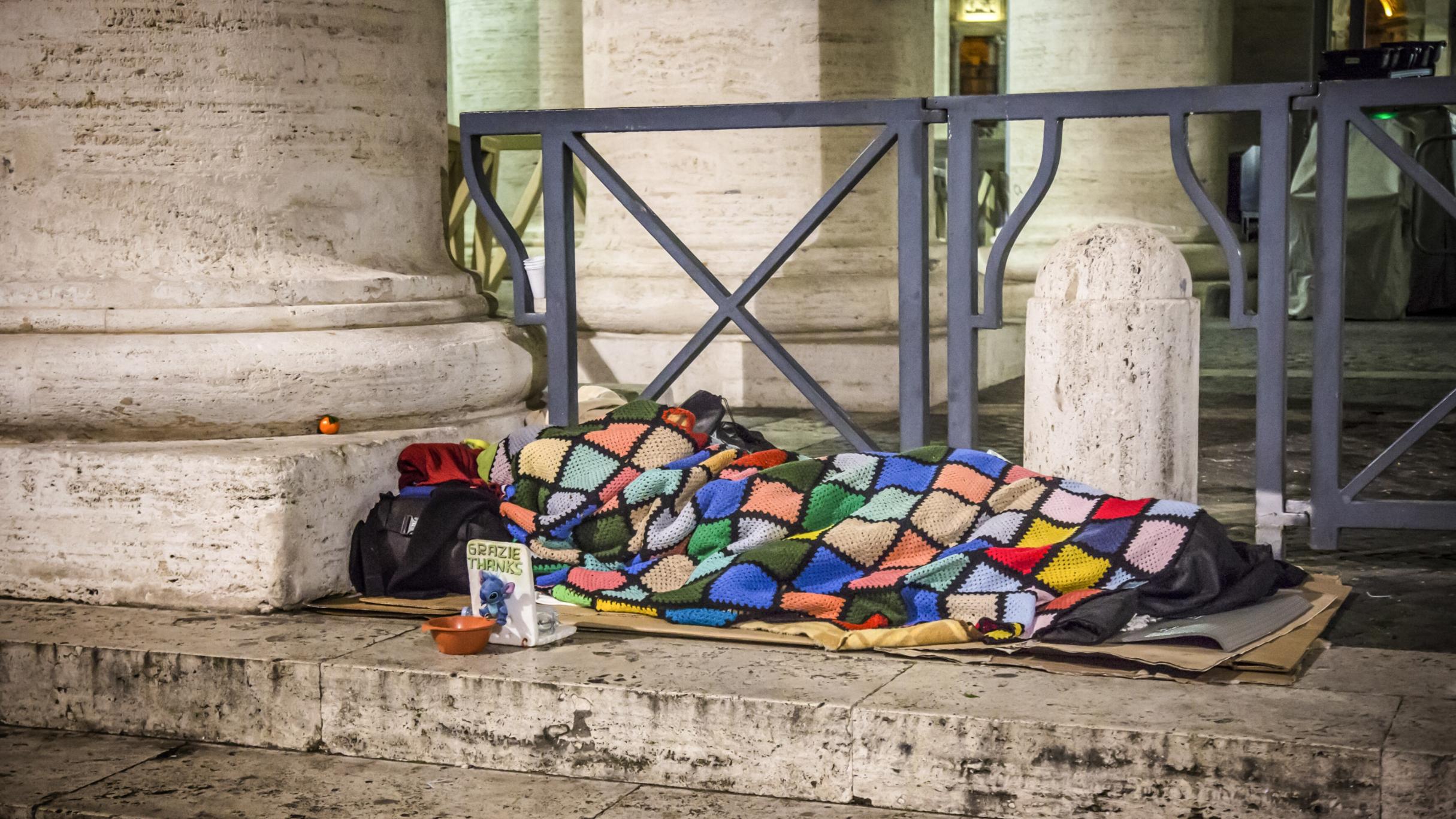 Ein Obdachloser schläft am 10. Januar 2017 auf Pappkartons unter einer bunten Decke auf dem Boden unter den Bernini-Kolonnaden am Petersplatz im Vatikan. Vor ihm steht ein Sammelbecher.