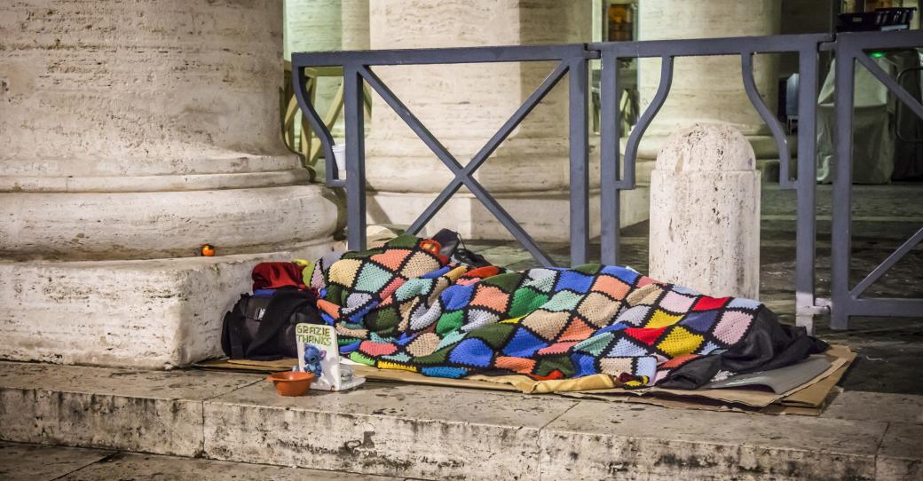 Ein Obdachloser schläft am 10. Januar 2017 auf Pappkartons unter einer bunten Decke auf dem Boden unter den Bernini-Kolonnaden am Petersplatz im Vatikan. Vor ihm steht ein Sammelbecher.