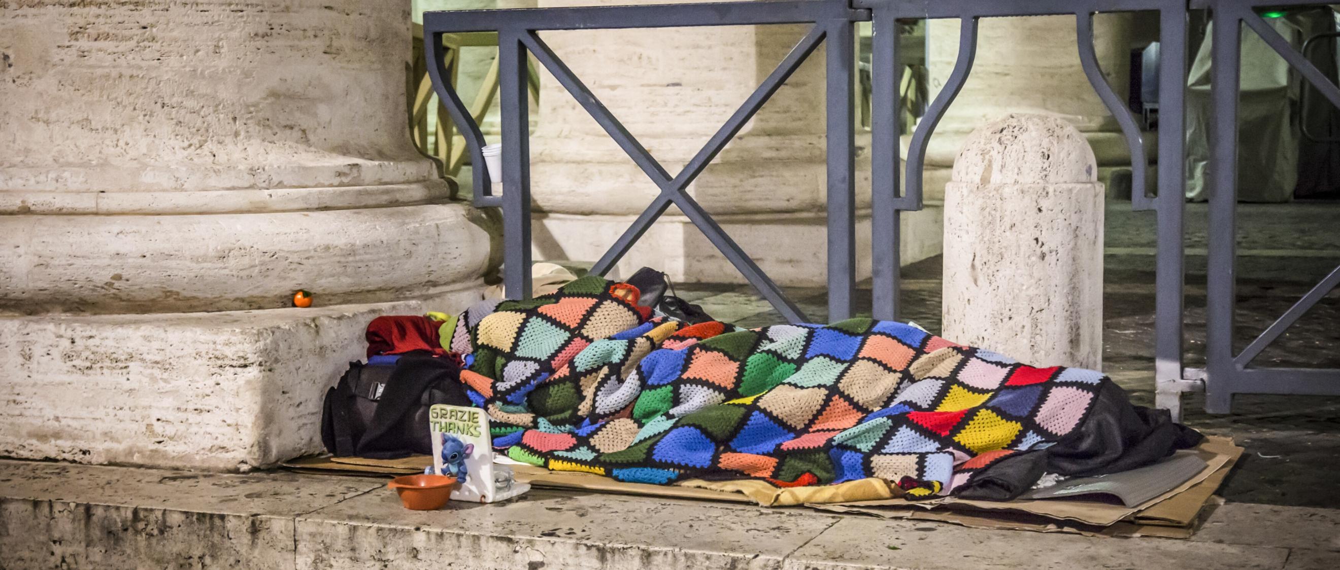 Ein Obdachloser schläft am 10. Januar 2017 auf Pappkartons unter einer bunten Decke auf dem Boden unter den Bernini-Kolonnaden am Petersplatz im Vatikan. Vor ihm steht ein Sammelbecher.