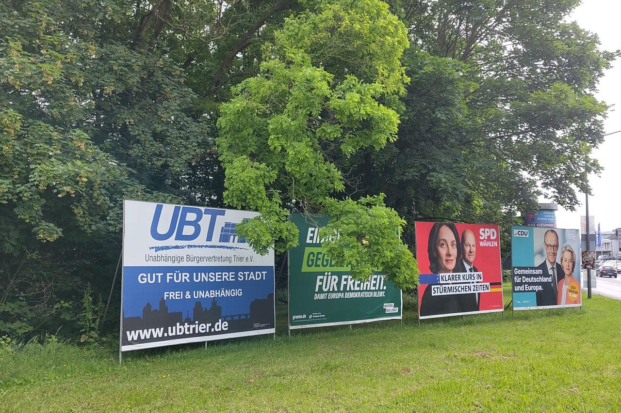 Das Foto zeigt vier Plakatwände mit Wahlplakaten zur Kommunal- und Europawahl, die nebeneinander auf einer Wiese am Rand einer Straße stehen; dahinter Bäume. Die Plakate (von links nach rechts) werben für die Unabhängige Bürgervertretung Trier (UBT), die Grünen, die SPD und die CDU.