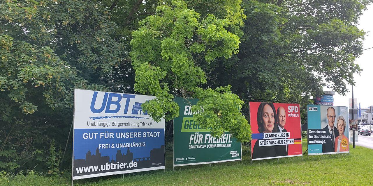 Das Foto zeigt vier Plakatwände mit Wahlplakaten zur Kommunal- und Europawahl, die nebeneinander auf einer Wiese am Rand einer Straße stehen; dahinter Bäume. Die Plakate (von links nach rechts) werben für die Unabhängige Bürgervertretung Trier (UBT), die Grünen, die SPD und die CDU.
