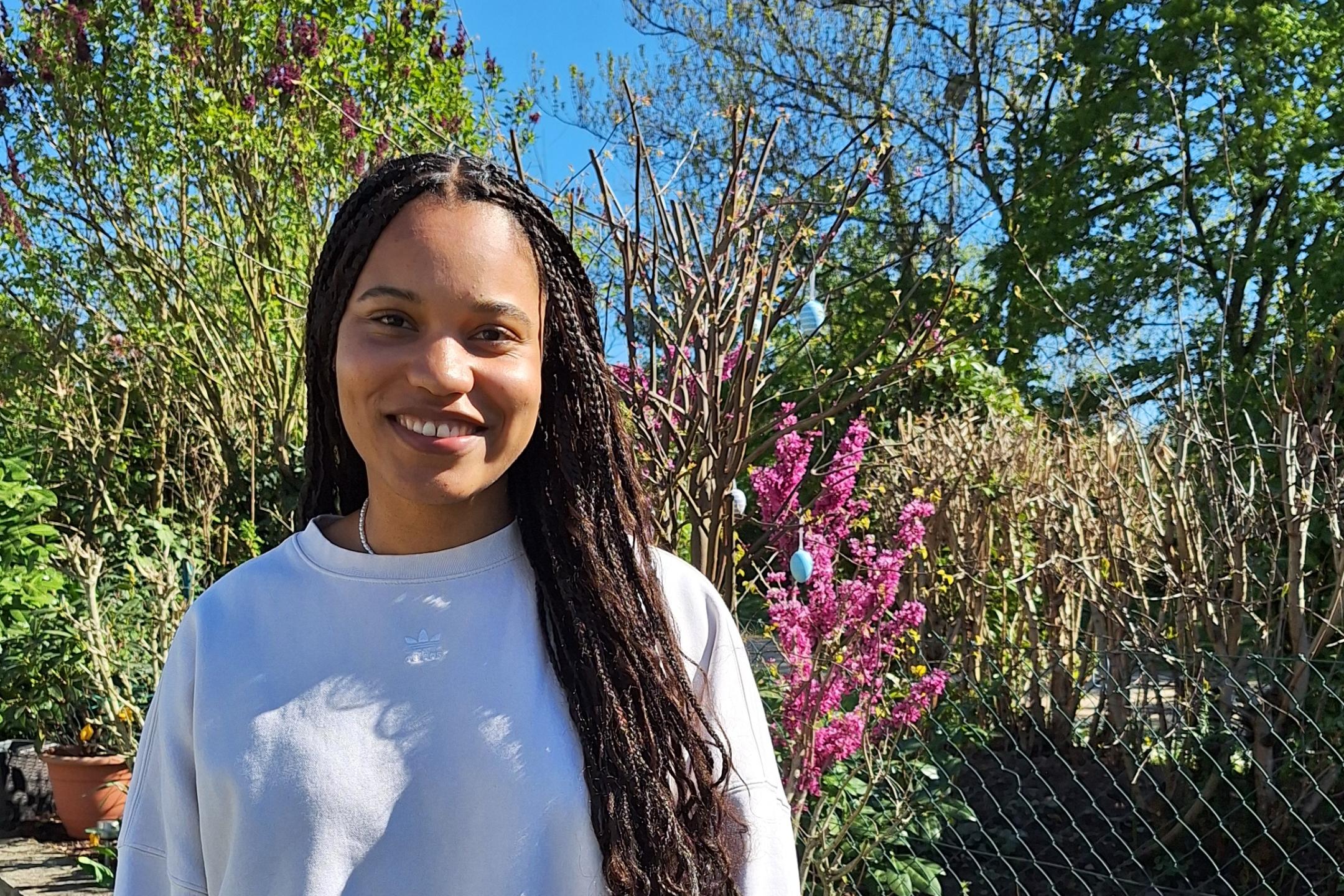 Eine junge Frau mit langen, geflochtenen Haaren – es handelt sich um die Kugelstoßerin Yemisi Ogunleye - steht lächelnd in einem sonnigen Garten vor einem Beet mit bunten Frühlingsblumen, darunter Tulpen und Hyazinthen. Sie trägt ein helles Sweatshirt und eine weiße Hose. Im Hintergrund sind blühende Sträucher, Bäume und ein blauer Himmel zu sehen.