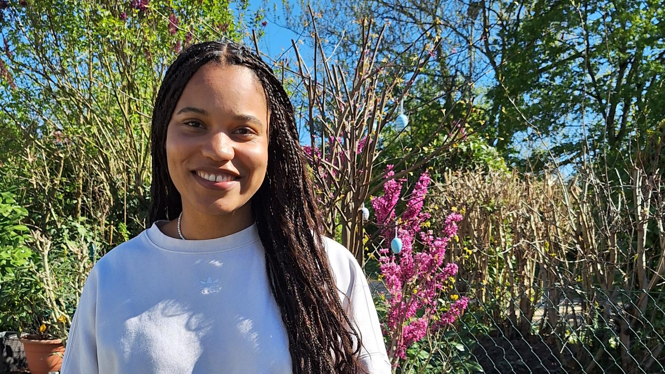 Eine junge Frau mit langen, geflochtenen Haaren – es handelt sich um die Kugelstoßerin Yemisi Ogunleye - steht lächelnd in einem sonnigen Garten vor einem Beet mit bunten Frühlingsblumen, darunter Tulpen und Hyazinthen. Sie trägt ein helles Sweatshirt und eine weiße Hose. Im Hintergrund sind blühende Sträucher, Bäume und ein blauer Himmel zu sehen.