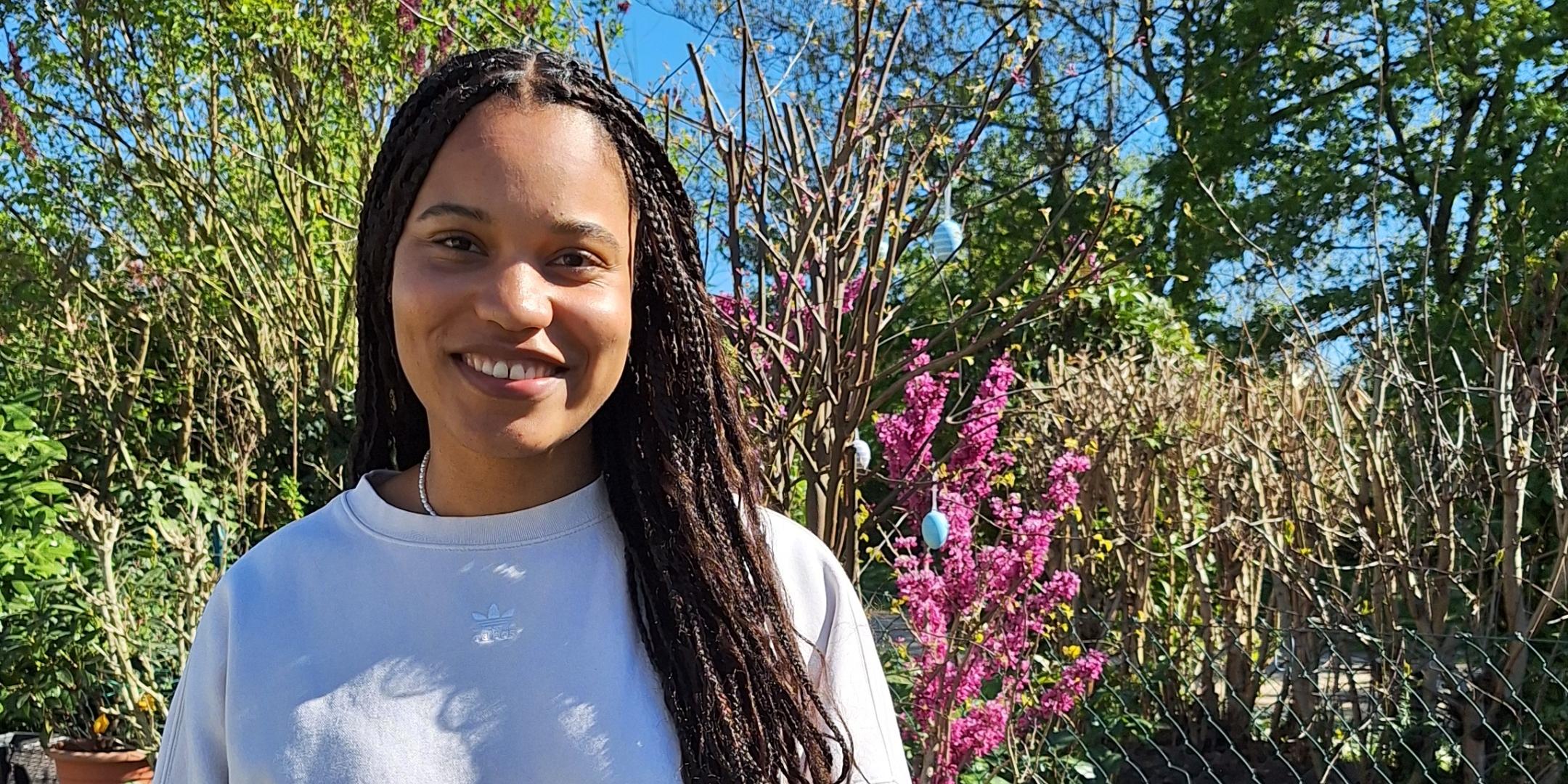 Eine junge Frau mit langen, geflochtenen Haaren – es handelt sich um die Kugelstoßerin Yemisi Ogunleye - steht lächelnd in einem sonnigen Garten vor einem Beet mit bunten Frühlingsblumen, darunter Tulpen und Hyazinthen. Sie trägt ein helles Sweatshirt und eine weiße Hose. Im Hintergrund sind blühende Sträucher, Bäume und ein blauer Himmel zu sehen.