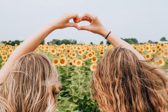 Abgebildet sind zwei Freundinnen in einem Sonnenblumenfeld, die mit ihren Händen ein Herz formen.