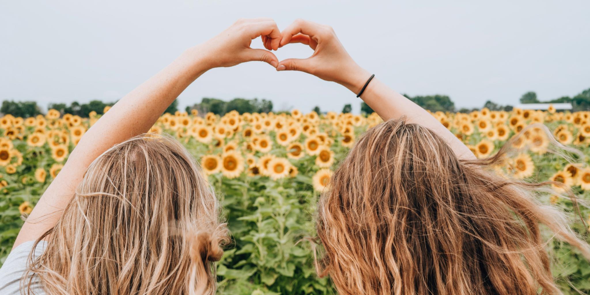 Abgebildet sind zwei Freundinnen in einem Sonnenblumenfeld, die mit ihren Händen ein Herz formen.