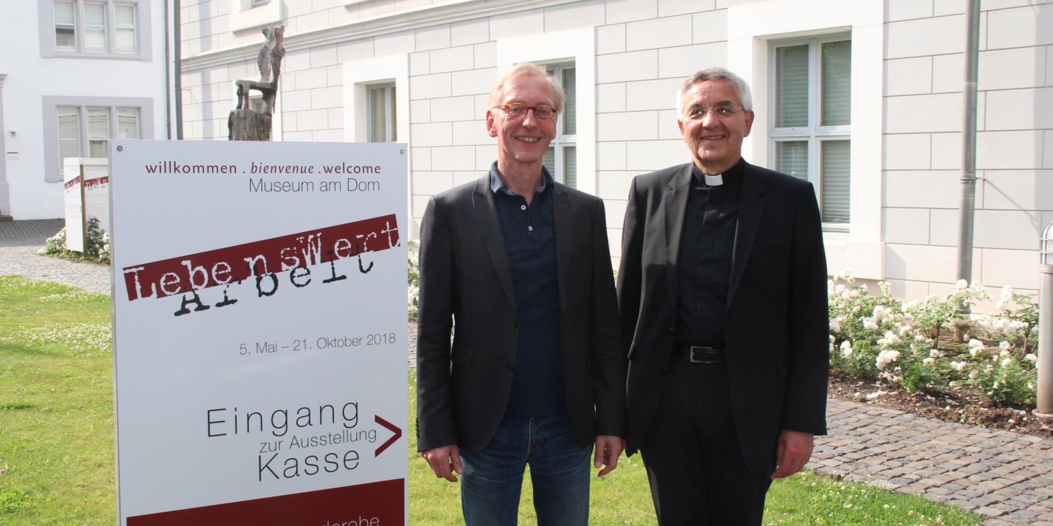 Professor Dr. Uwe Becker (links) und Weihbischof Franz Josef Gebert vor dem Ausstellungsplakat 'LebensWertArbeit'. Foto: DiCV Trier