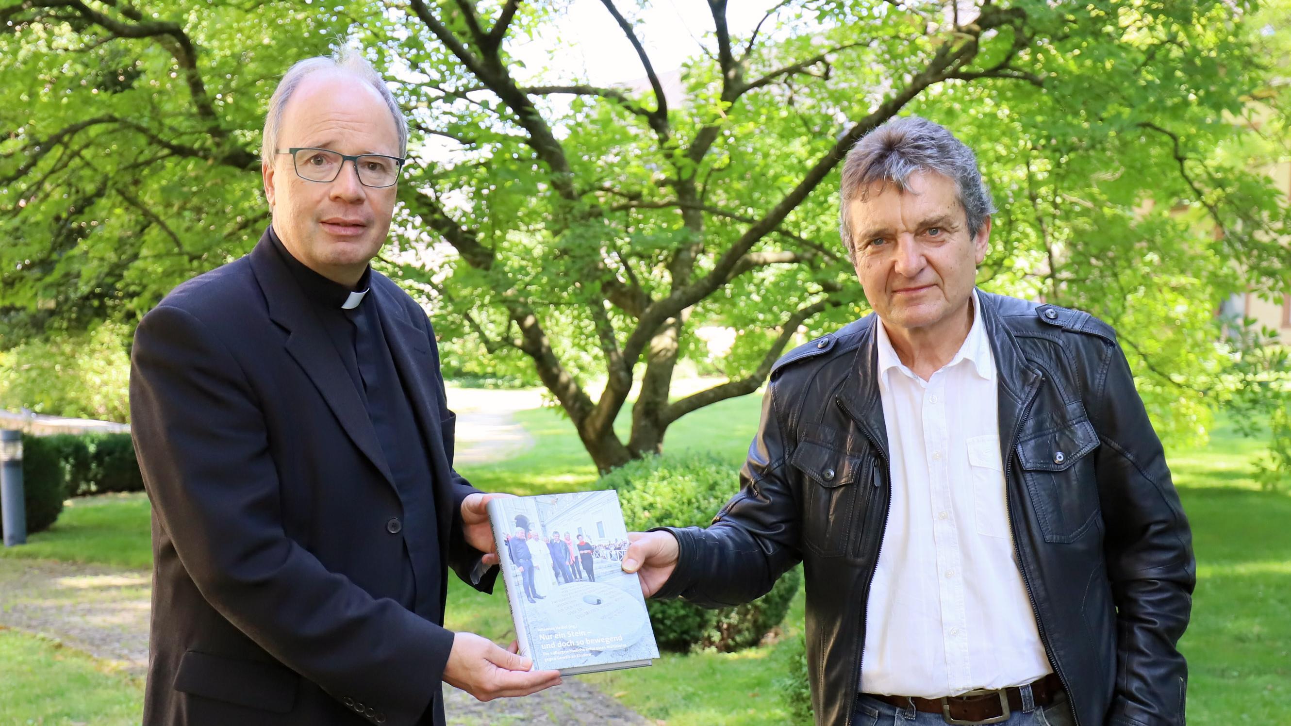 Johannes Heibel (links) übergibt den Bildband zum „Mahnenden Mühlstein“ an Bischof Ackermann. (Foto: Christina Libeaux/Bistum Trier)