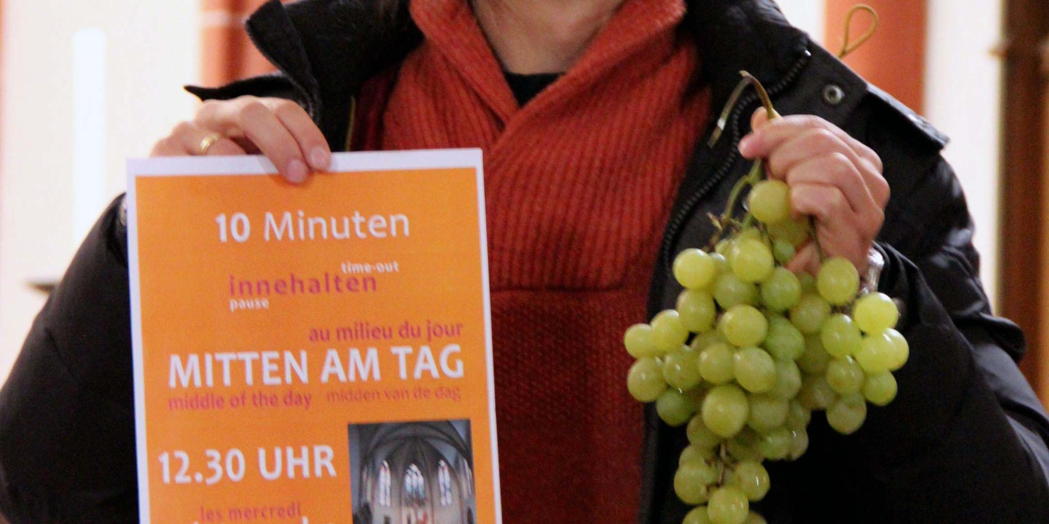 Pastoralreferent Tomek Welke zeigt das Plakat 'Mitten am Tag' und die für die Region typischen Weintrauben