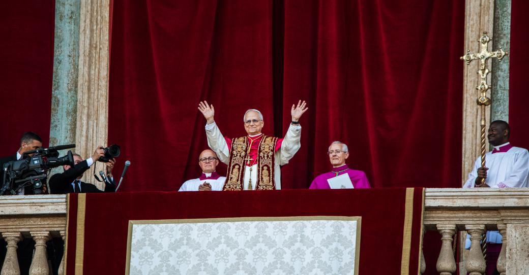 Der Papst steht auf dem Balkon des Petersdom und hebt beide Hände, zu seinem beiden Seiten sitzen zwei Kardinäle, hinter ihm ein roter Samtvorhang, an der Seite hängt ein Kreuz.