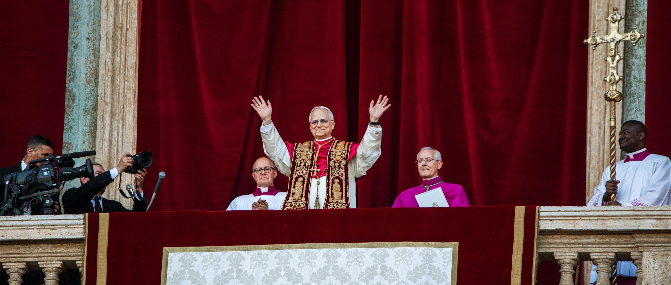 Der Papst steht auf dem Balkon des Petersdom und hebt beide Hände, zu seinem beiden Seiten sitzen zwei Kardinäle, hinter ihm ein roter Samtvorhang, an der Seite hängt ein Kreuz.