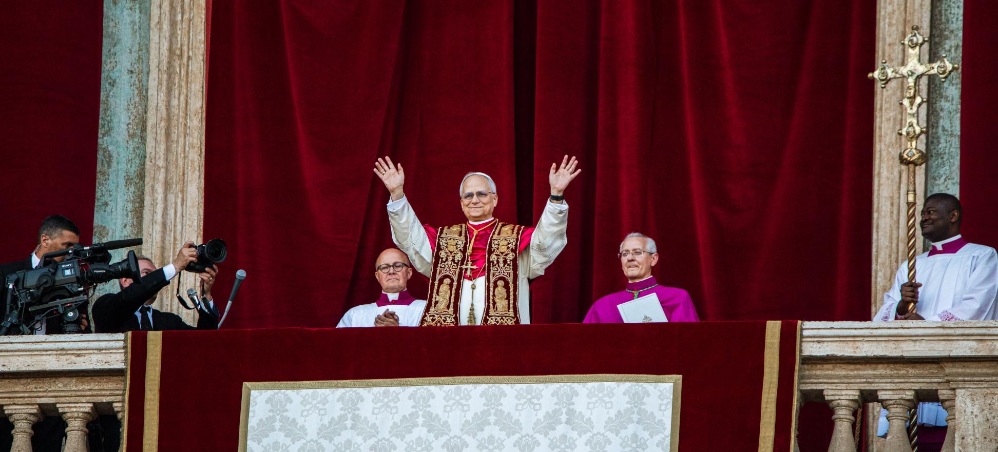 Der Papst steht auf dem Balkon des Petersdom und hebt beide Hände, zu seinem beiden Seiten sitzen zwei Kardinäle, hinter ihm ein roter Samtvorhang, an der Seite hängt ein Kreuz.