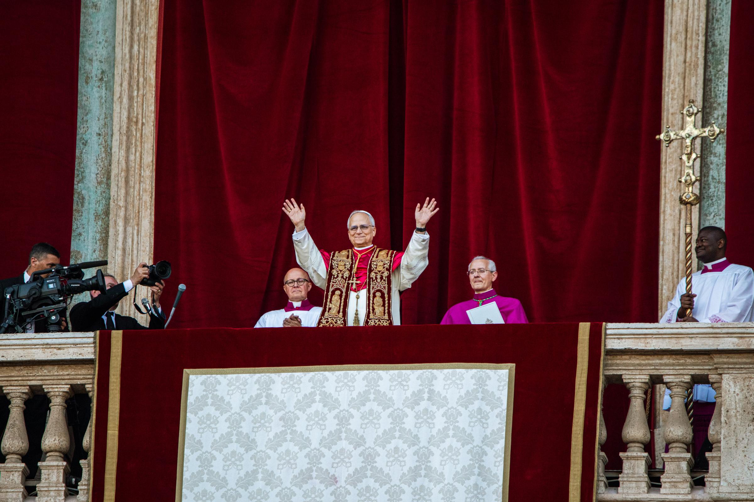 Der Papst steht auf dem Balkon des Petersdom und hebt beide Hände, zu seinem beiden Seiten sitzen zwei Kardinäle, hinter ihm ein roter Samtvorhang, an der Seite hängt ein Kreuz.