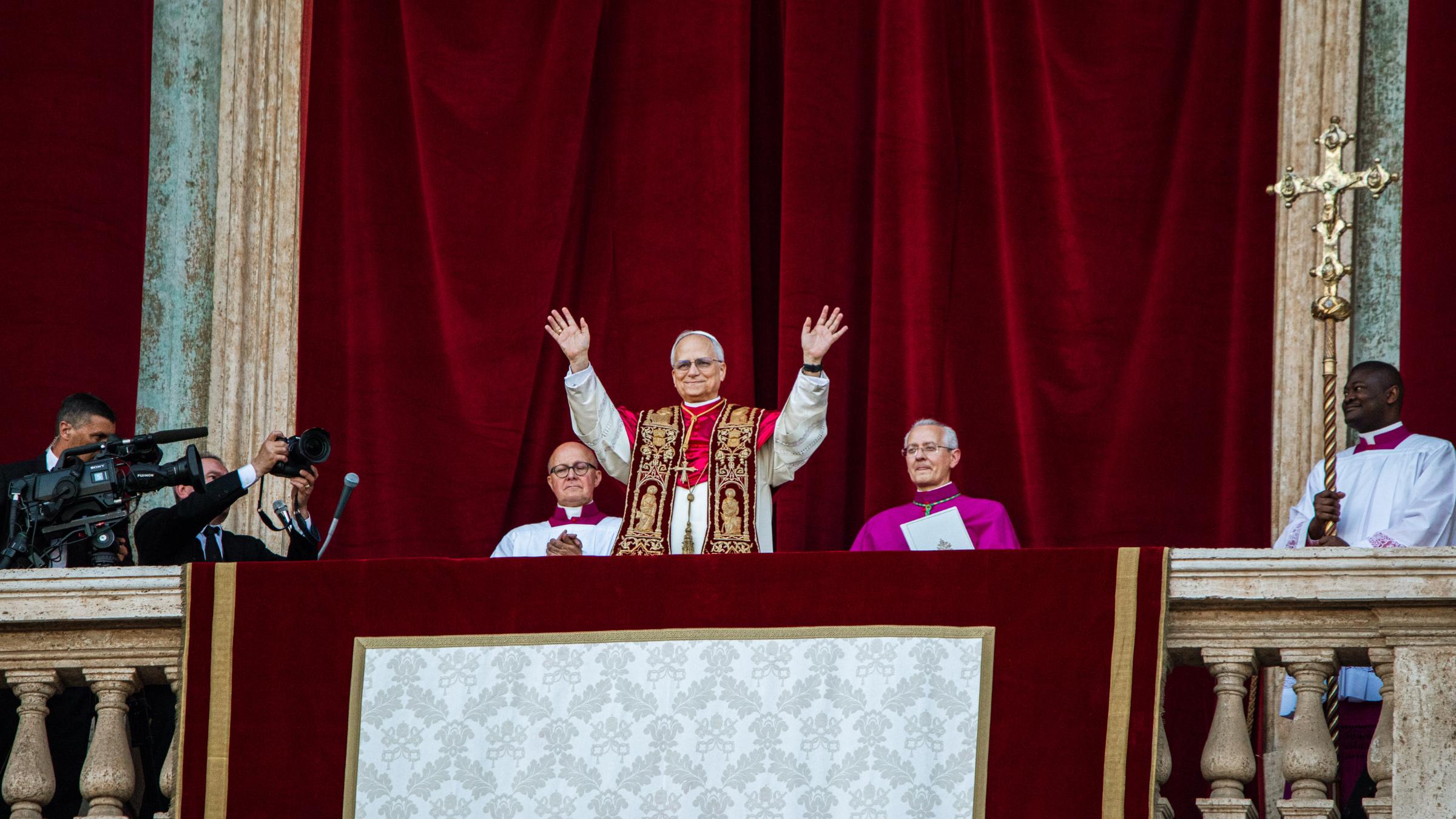 Der Papst steht auf dem Balkon des Petersdom und hebt beide Hände, zu seinem beiden Seiten sitzen zwei Kardinäle, hinter ihm ein roter Samtvorhang, an der Seite hängt ein Kreuz.