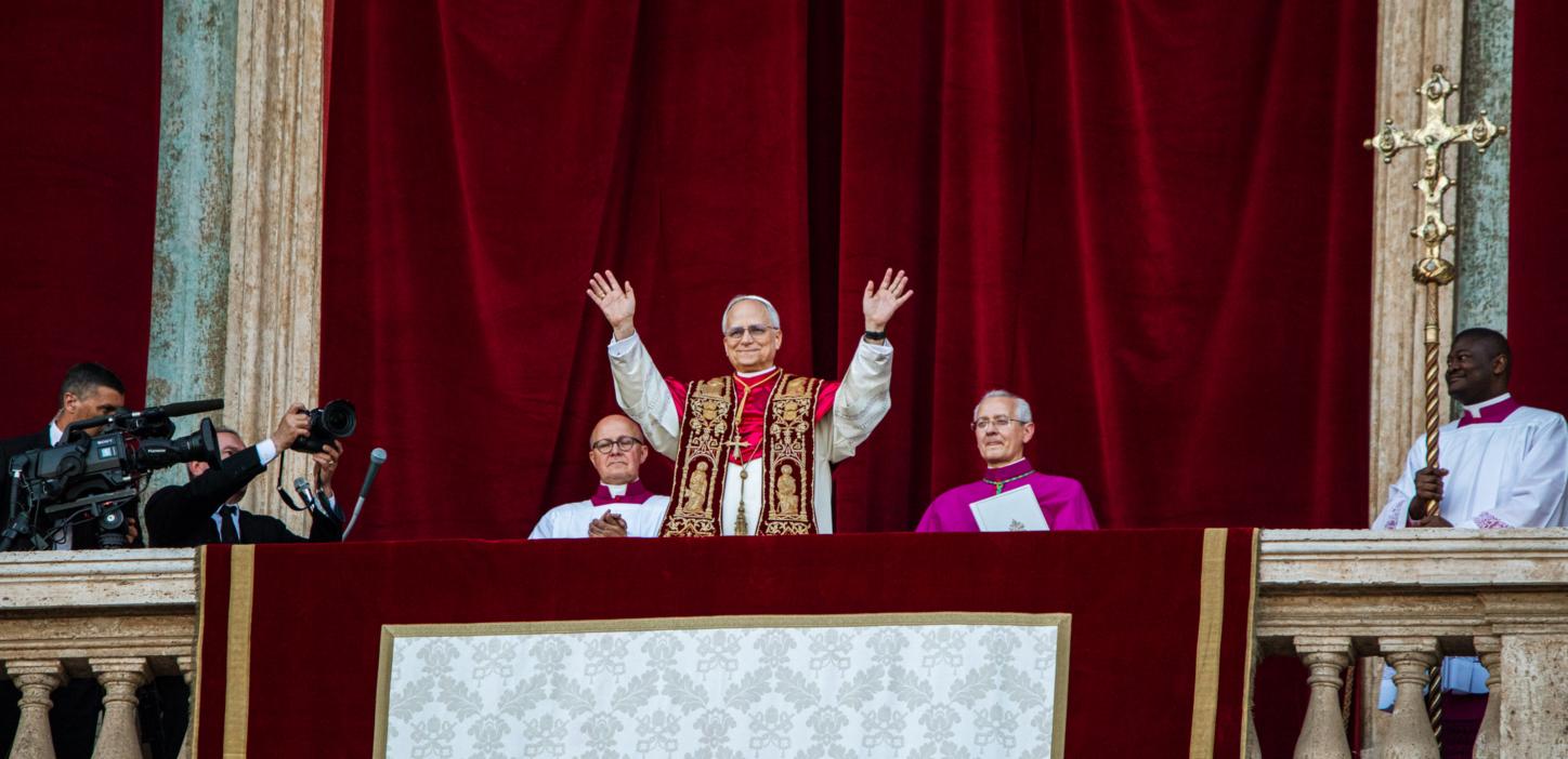 Der Papst steht auf dem Balkon des Petersdom und hebt beide Hände, zu seinem beiden Seiten sitzen zwei Kardinäle, hinter ihm ein roter Samtvorhang, an der Seite hängt ein Kreuz.